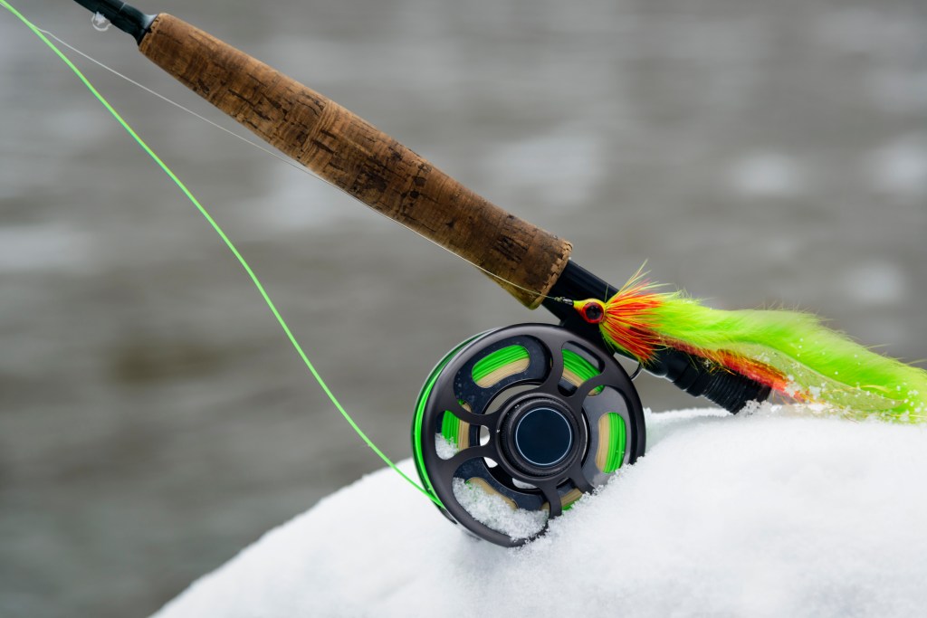 Fly fishing rod and bait sit on snow in front of a blurred river background.