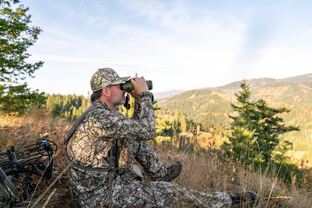 A bowhunter sits on a mountain peak and looks through binoculars while tracking wild game in the forested wilderness of Washington State. A crossbow is lying on the ground behind the man.