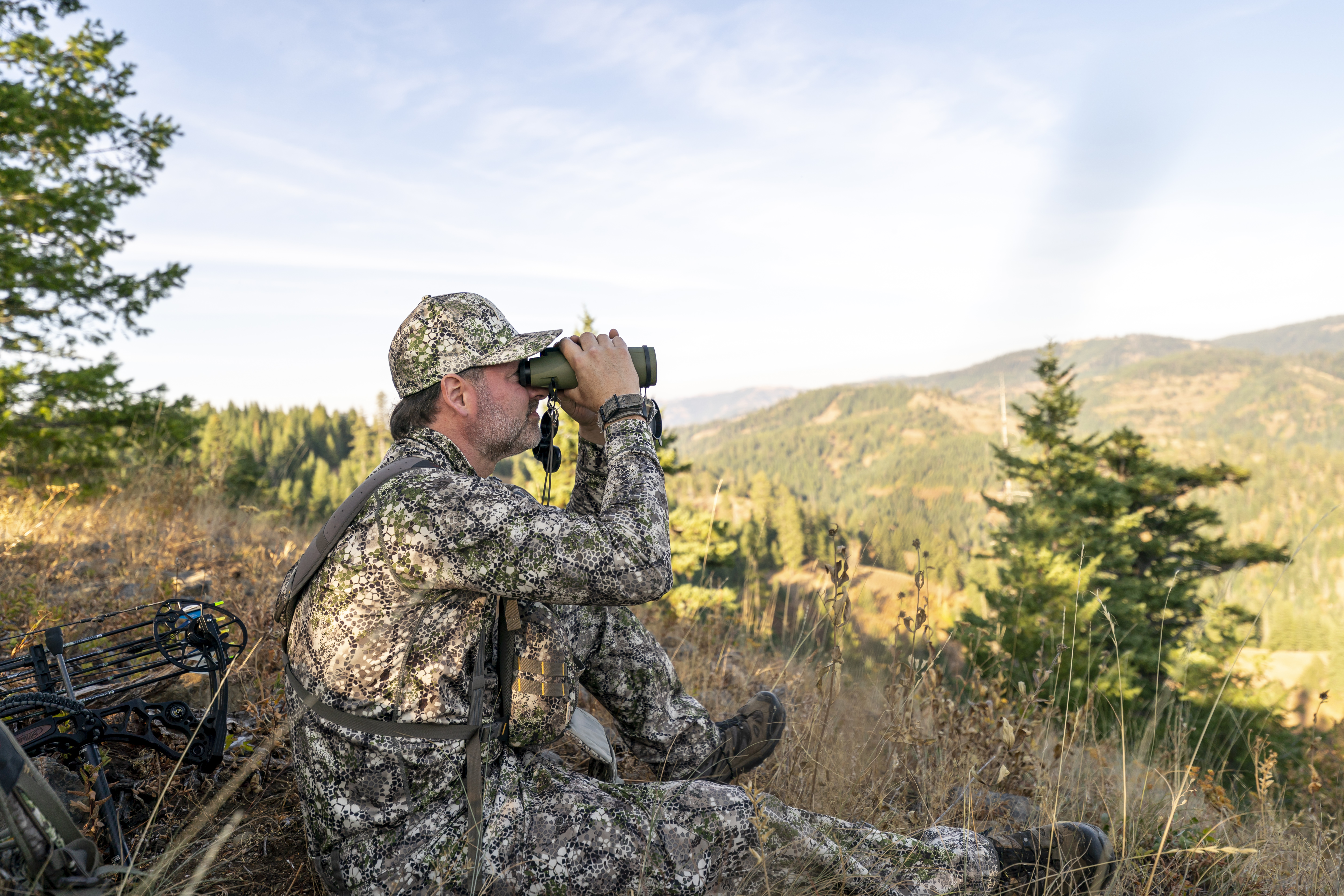 A bowhunter sits on a mountain peak and looks through binoculars while tracking wild game in the forested wilderness of Washington State. A crossbow is lying on the ground behind the man.