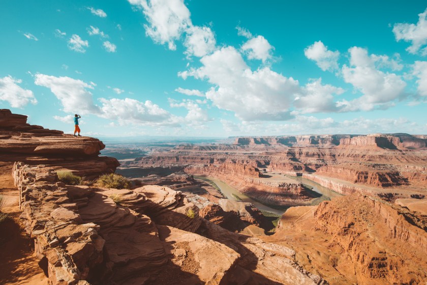 A young male hiker is standing on the edge of a cliff enjoying a dramatic overlook of the famous Colorado River and beautiful Canyonlands National Park in scenic Dead Horse Point State Park, Utah, USA