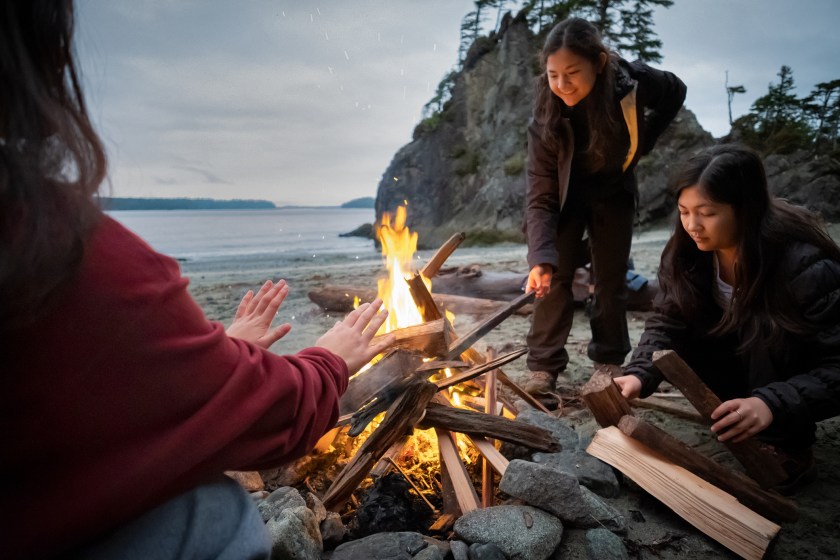 Eurasian Generation Z sisters in winter clothing add kindling to campfire. Pacific Ocean in background. Bamfield, Vancouver Island, British Columbia, Canada