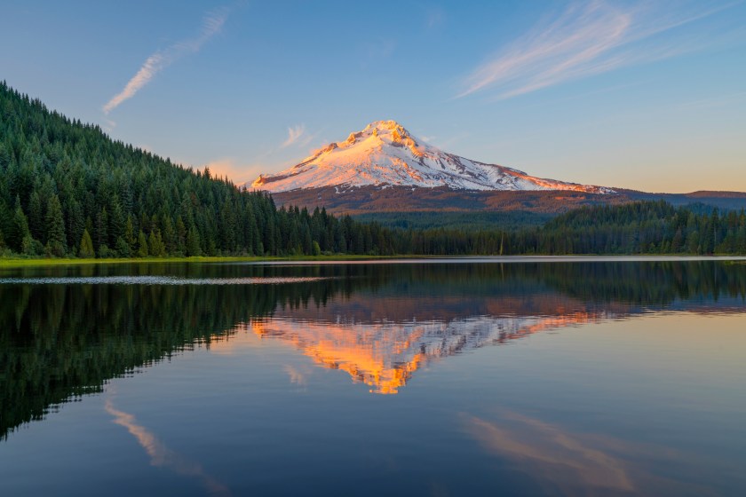 Trillium Lake in Oregon at the water's edge of the lake to see golden light on Mount Hood peak during sunset. Mount Hood mountain had golden light shining on the peak at sunset. A beautiful sunset adventure at Trillium lake near Portland, Oregon in the Pacific Northwest after a hike. Reflections of Mt Hood were seen in Trillium Lake water with trees around the lake. There were amazing clouds in the sky during sunset at Trillium lake with a view of Mount Hood.