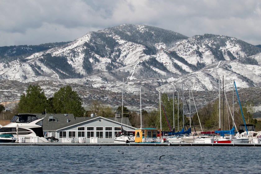 With the snow covered Rocky Mountains and green trees in the background, sail and motor boats remain docked at the marina at Chatfield Reservoir State Park in Colorado.