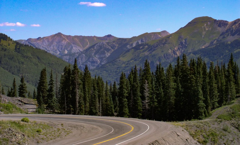View of a paved cure in the Million Dollar Highway in Colorado with trees and layers of mountains in the background.