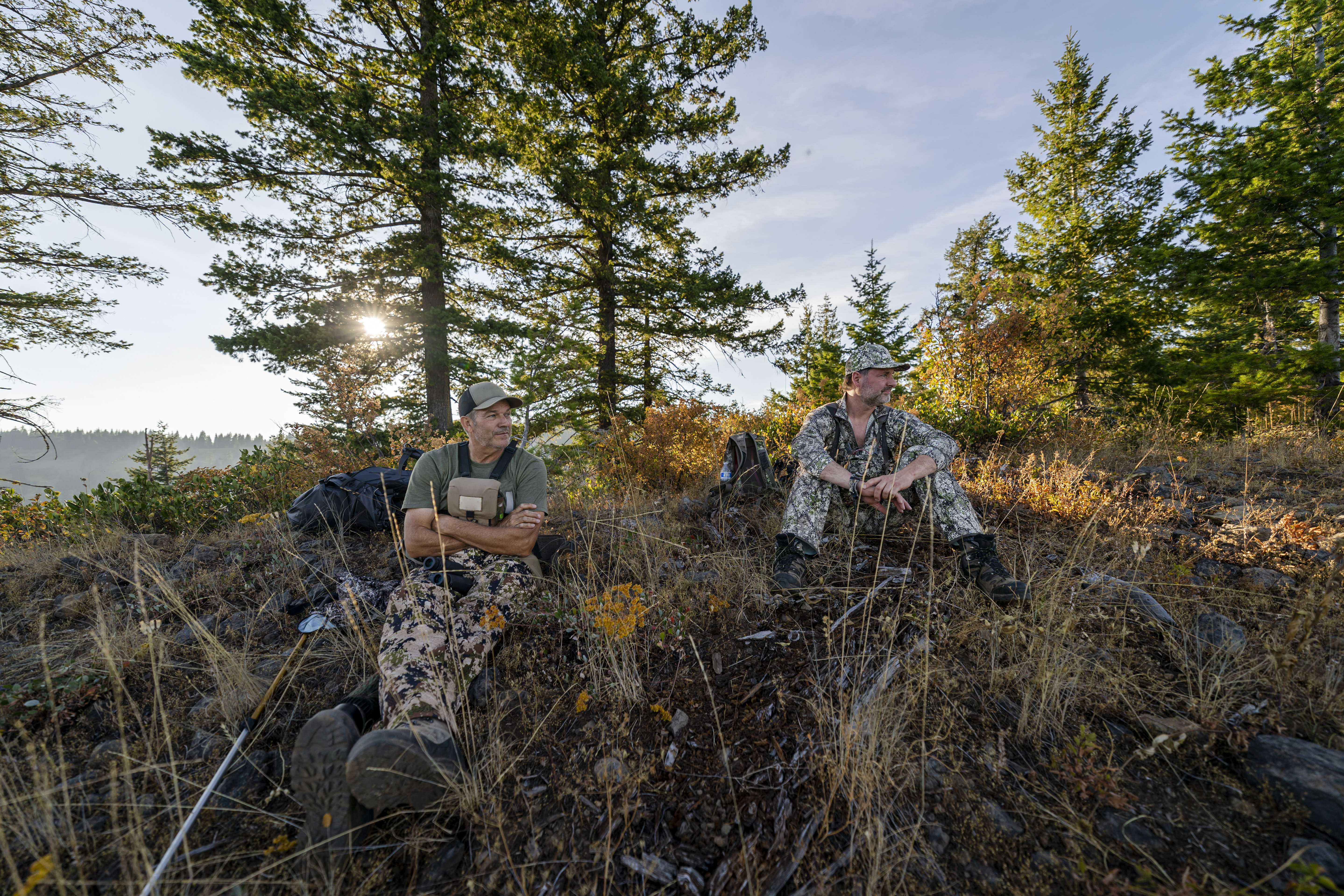 Two middle aged men wearing camouflage clothing sit on a mountainside in Washington state while hunting elk with a crossbow. It is a warm and sunny Autumn afternoon.