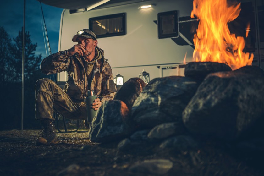 Caucasian Hunter in His 40s Wearing Camouflage Drinking Hot Drink Next to His Motorhome and Campfire. Hunting Season and Outdoors Theme.