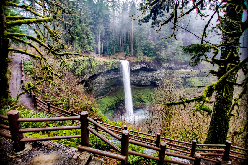 A view of South Falls, a massive rushing waterfall surrounded by lush forests and wooden fence in foreground