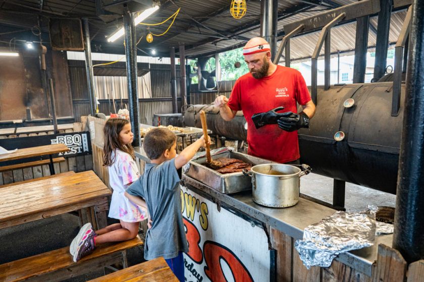 LEXINGTON, TEXAS - JULY 31: Pitmaster Clay Cowgill helps two young patrons try their hand at mopping the ribs at Snow's BBQ on July 31, 2021 in Lexington, Texas