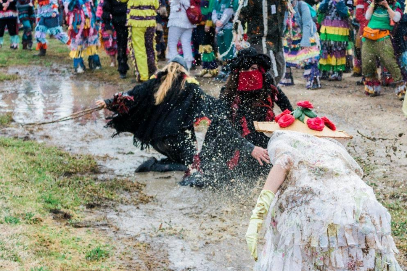 three brightly costumed people chase each other in the mud as a crowd watches on