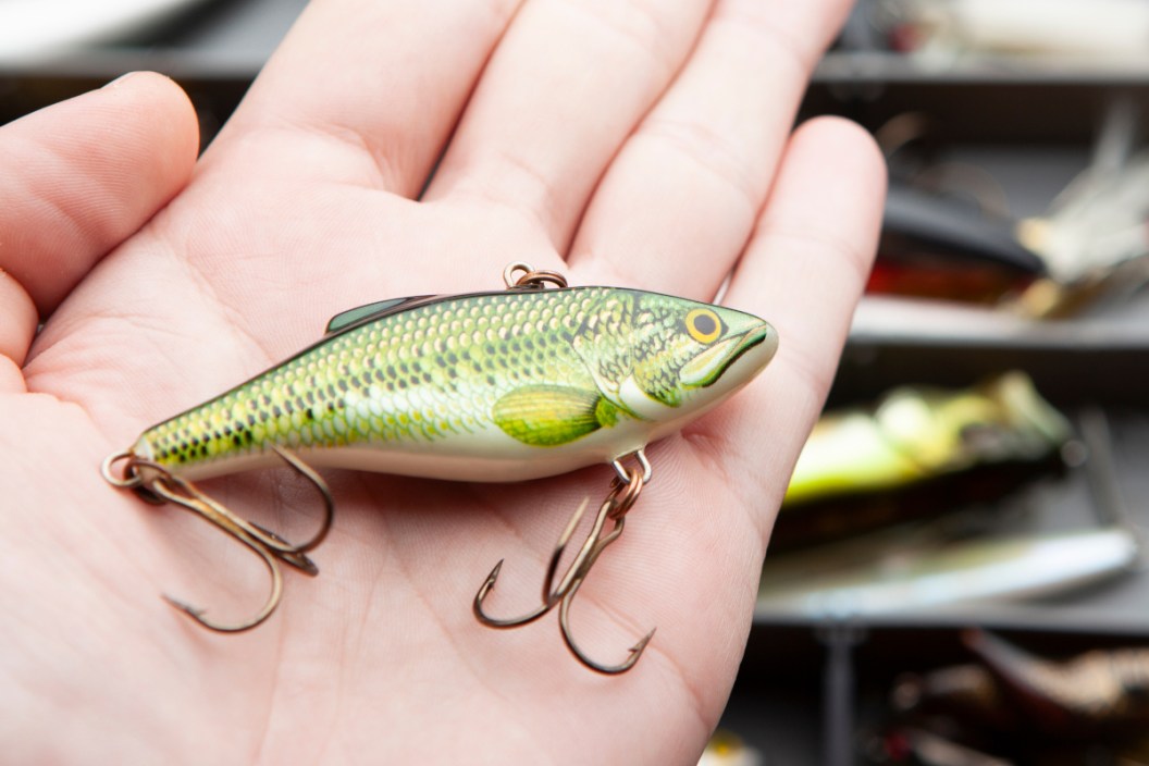 A hand holding a lipless crankbait above a tackle box.