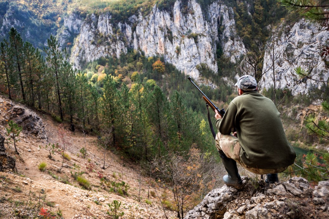 Portrait of hunter with rifle crouching on top of the hill scouting his game