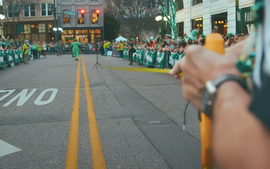 a man holds out a tape measure while a man dressed as a leprechaun measures a street while people in green watch on