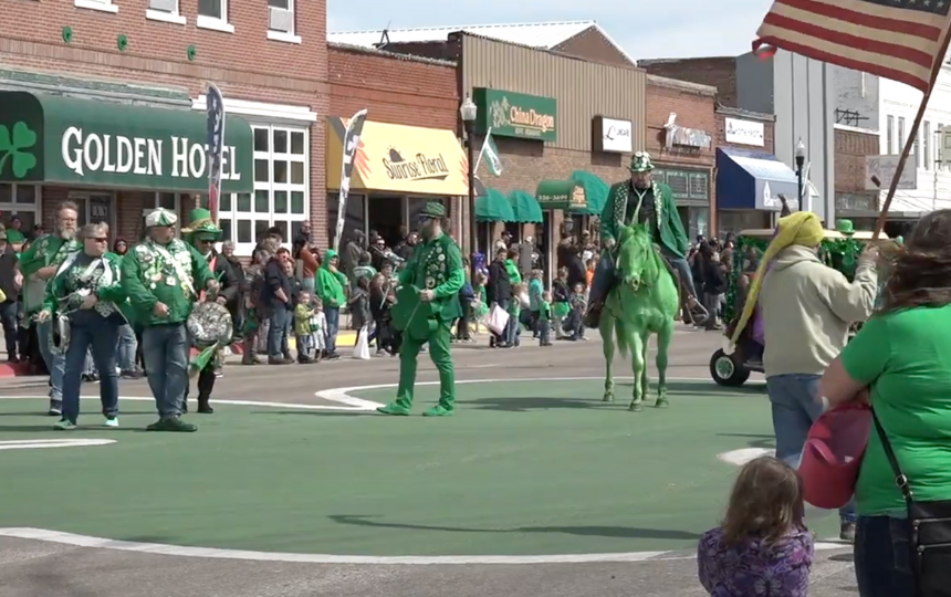 a St. Patrick's Day parade in a small town main street; men in green riding green horses ride over a giant green shamrock painted on the street