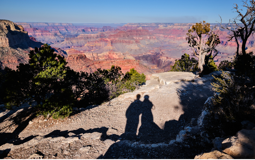 Shadows of two people touching at the shoulder on outcrop near Yavapai Point with a wide view of Grand Canyon spreading out in the background
