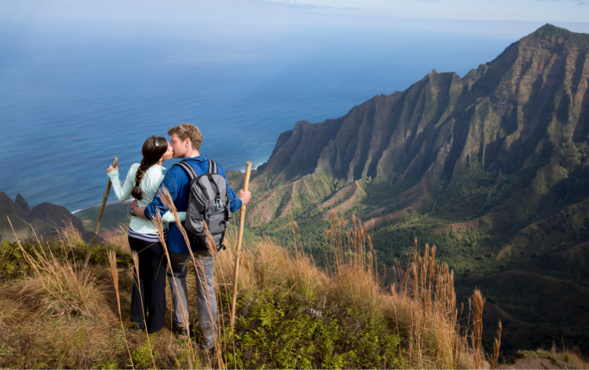 A husband and wife are hiking on their honeymoon in the Hawaiian mountains. They are kissing on the ridge overlooking a beautiful view.