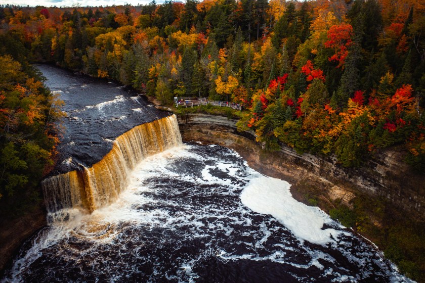 Close up aerial photograph of the upper waterfall cascade at Tahquamenon Falls with beautiful autumn colored foliage of green, yellow, red and orange tree leaves as tourists line the platform below