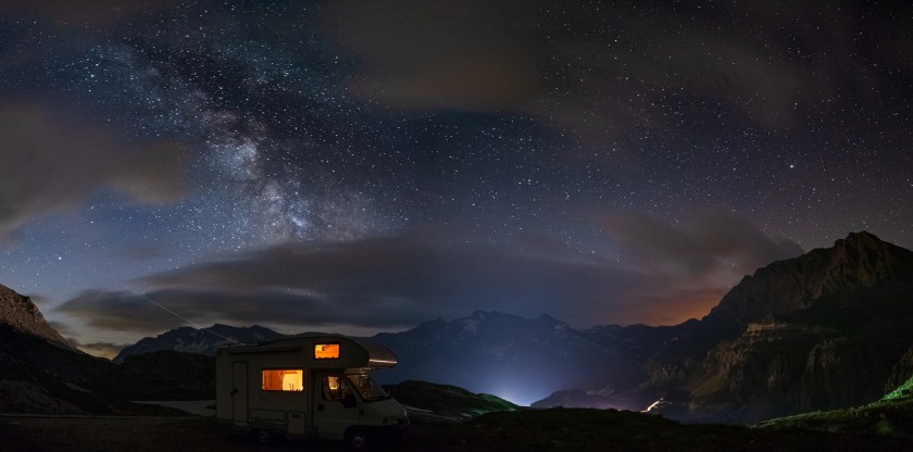 Panoramic night sky over the Alps. The Milky Way galaxy arc and stars over illuminated camper van. Camping freedom in unique landscape.
