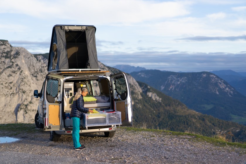 Woman Standing At Back of Her Camper Van with Open Rooftop Tent, Indoor Bed and Drawer with Boxes high up in the Mountains on her Roadtrip Through Austria