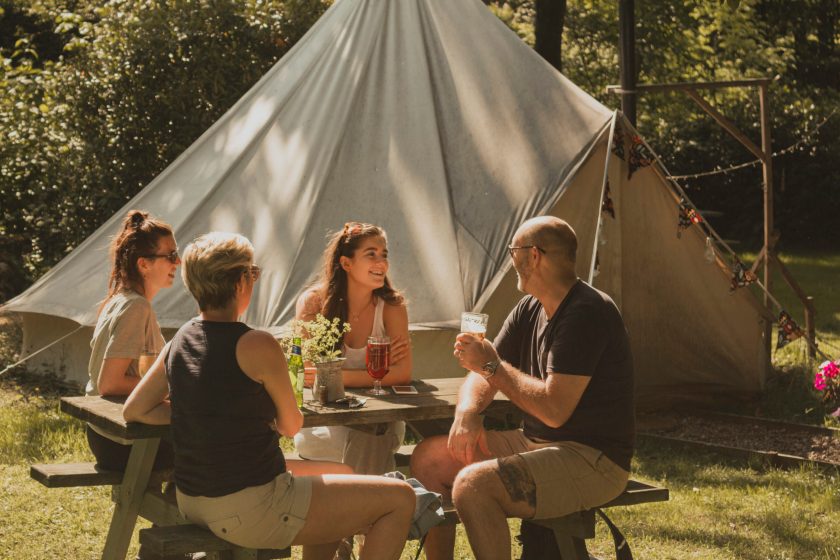 Family of four are sitting at a picnic bench next to their glamping tent. They are all talking and enjoying an alcoholic beverage.