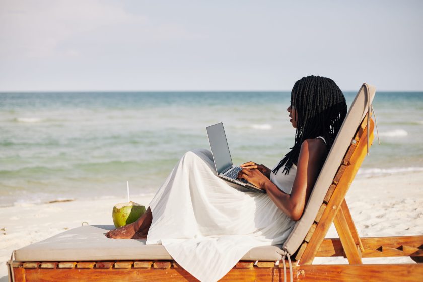 Young woman enjoying sitting on chaise-lounge, working on laptop and looking on sea waves