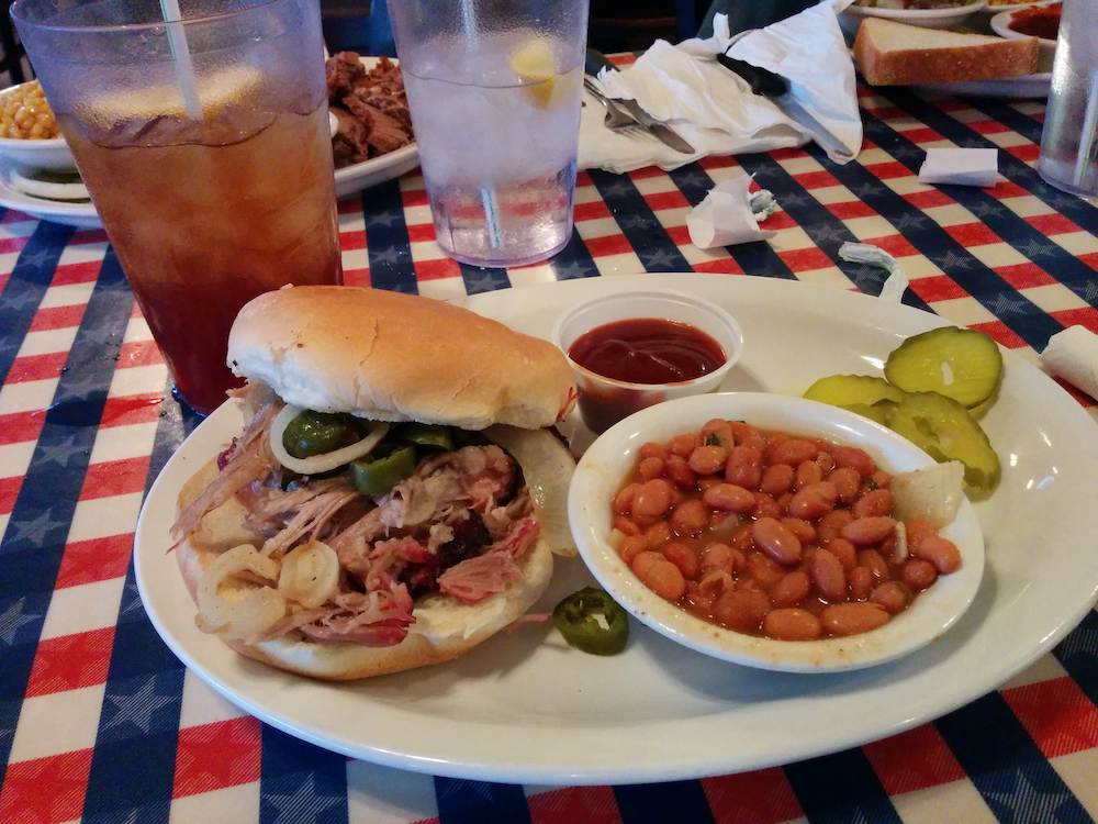 A delicious pulled pork sandwich with onions and jalapenos with a side of baked pinto beans on a white plate with some sweet tea at a traditional american barbecue BBQ restaurant near Houston, Texas, USA.