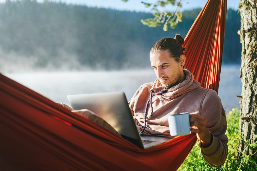 Young man in hammock working on his laptop and drinking hot coffee. Leisure activities / Remote working concept.