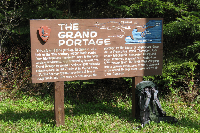 a large brown sign reads "THE GRAND PORTAGE: This 8.5 mile long portage became a vital link in the 18th century water trade route from Montreal and the Great Lakes to the north-western wilderness. First traveled by Indians, the Grand Portage by-passed a series of falls and rapids extending along the last 21 miles of the Pigeon River. During the fur-trade, thousands of tons of trade goods and furs were carried across the portage on the backs of voyageurs. Sieur de La Verendrye, David Thompson, Alexander MacKenzie, Alexander Henry and other explorers travled this route from 1729 through 1802. The North West Company held a summer rendevous at the eastern end of the Grand Portage along these shores of Lake Superior."