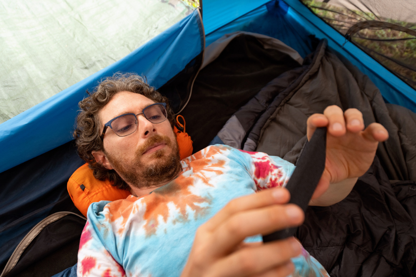 While tent camping a caucasian man in his 30s is wearing a tie dye t-shirt and spends time relaxing watching something on his mobile phone on a summer day in Hot Springs National Park Arkansas, USA.