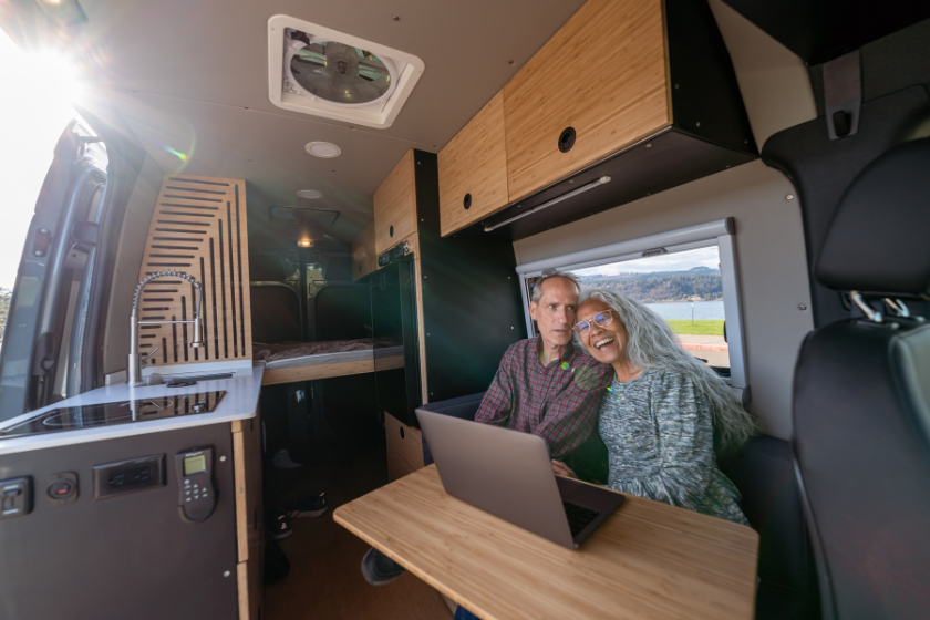An affectionate senior couple relax together in their customized camper van and enjoy the view of their campsite visible through the van's open door. A laptop computer is sitting on the table in front of the retired couple.