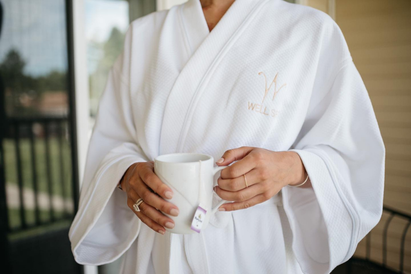 torso picture of a woman wearing a Wells Spa branded bathrobe holding a cup of tea