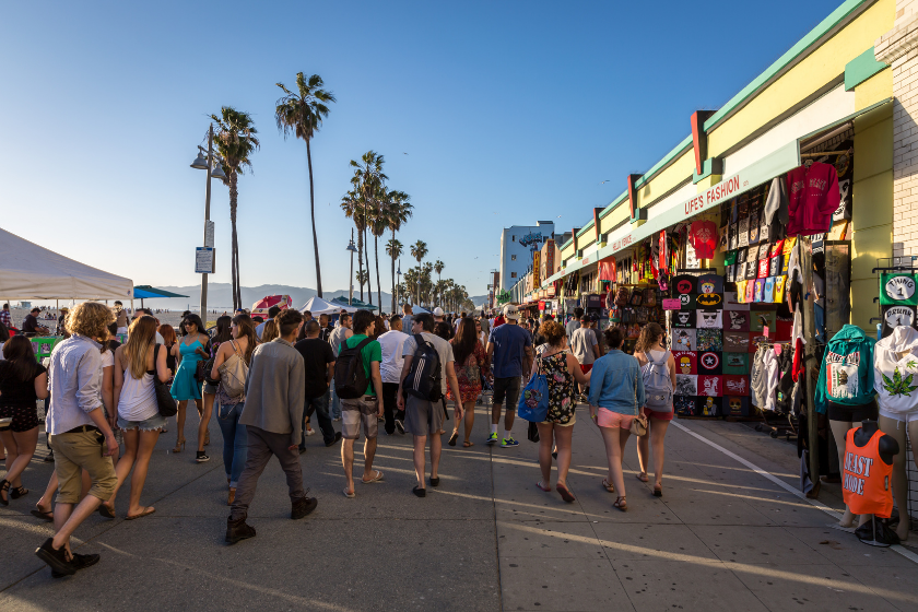 Tourists and locals are enjoying a sunny day along the famous Venice Beach promenade. The boardwalk features performers, vendors, and artists selling their craft. Bikers are riding on the curved bike-way along the boardwalk.