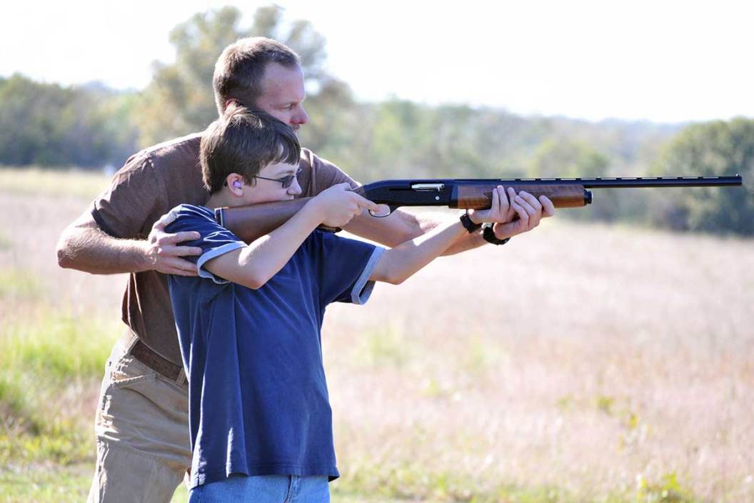 Older man helps show a young boy how to hold and shoot a shotgun.