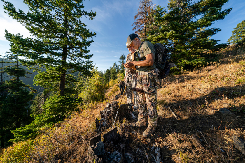 Two bowhunters tracking elk in a mountain range located in the Pacific Northwest region of the USA check their location using GPS on a mobile phone. One of the men is holding a bull elk grunt tube between his knees. The other man is holding a crossbow.