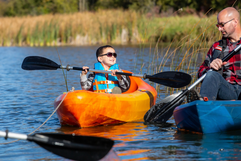 Close-up of a four year old boy having fun in his kayak on the lake. The boy is wearing sunglasses and a life jacket and is using a black paddle. His kayak is orange and yellow. In this image he is looking up towards dad.