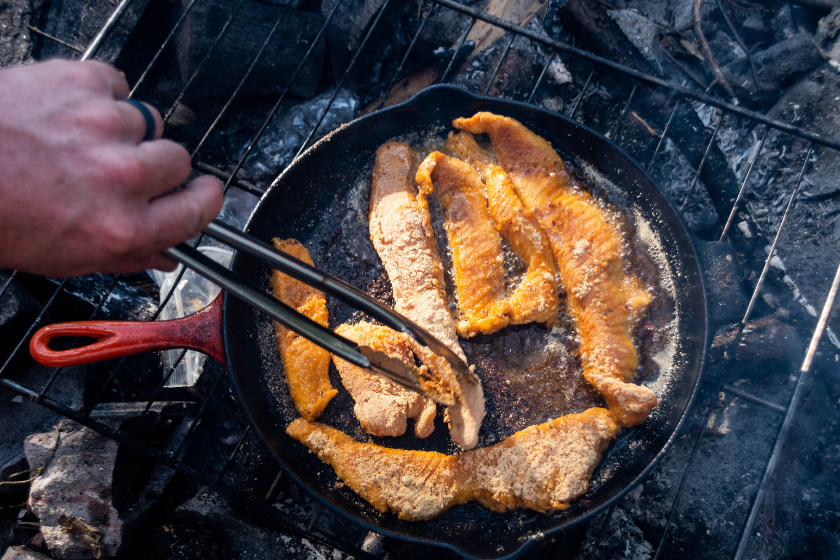 A meal of fish and sides frying on a cast iron pan on a campfire.