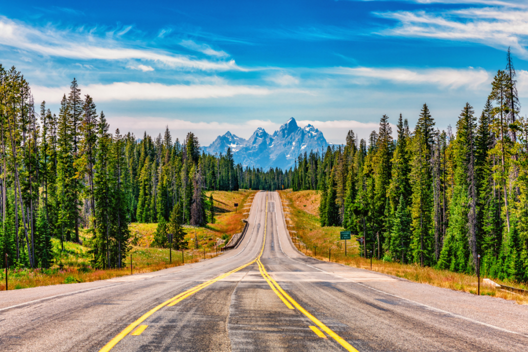 A mountain highway through the forest toward Grand Teton National Park, Wyoming with the peaks of the Tetons in the background.