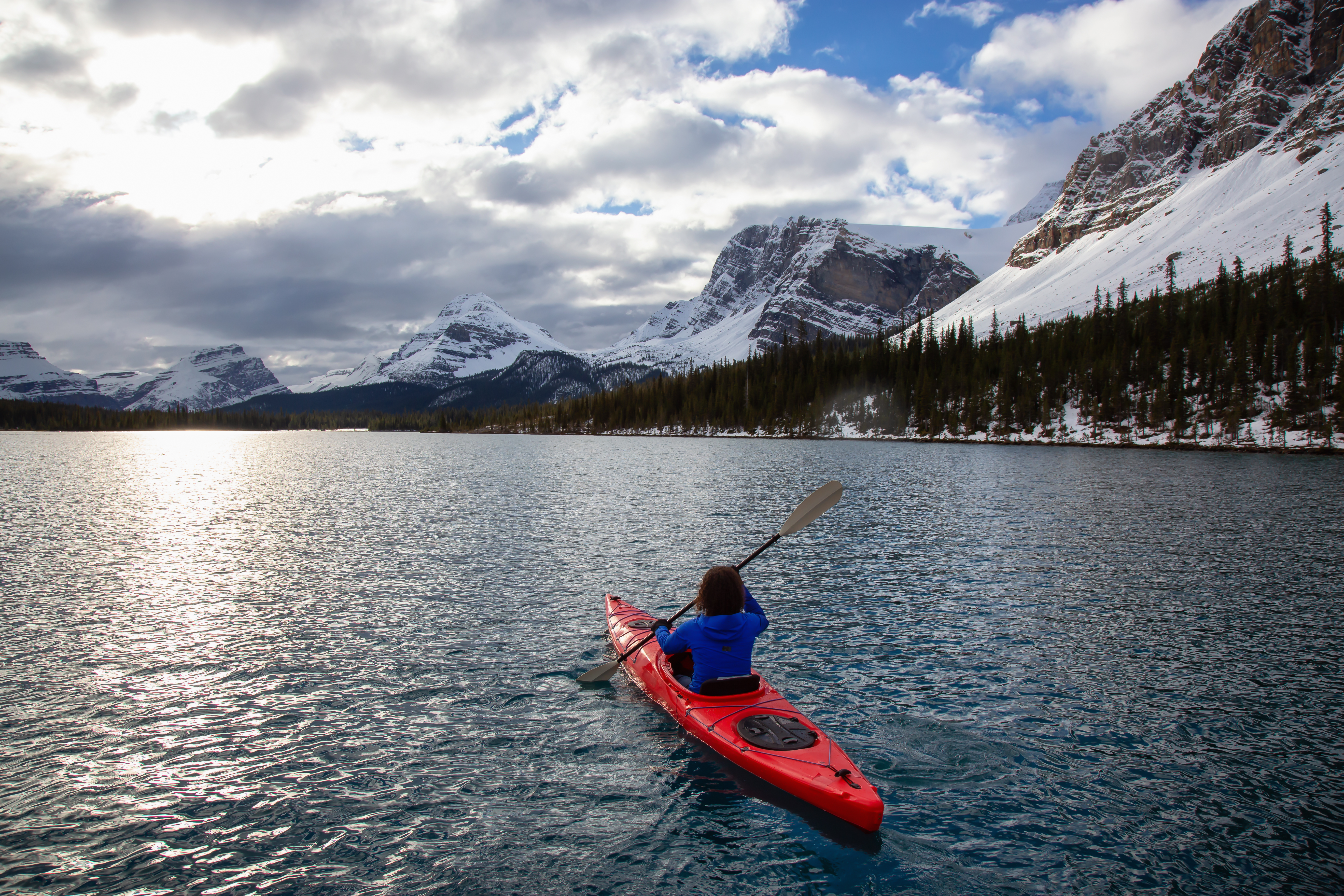 Adventurous girl kayaking in a glacier lake surrounded by the Canadian Rockies during a cloudy morning. Taken at Bow Lake, Banff, Alberta, Canada.