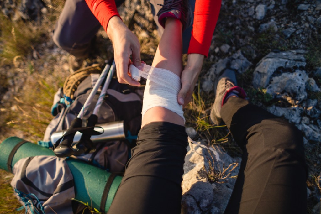 Young man with first aid kit wrapping an injured leg of a female hiker with a bandage.