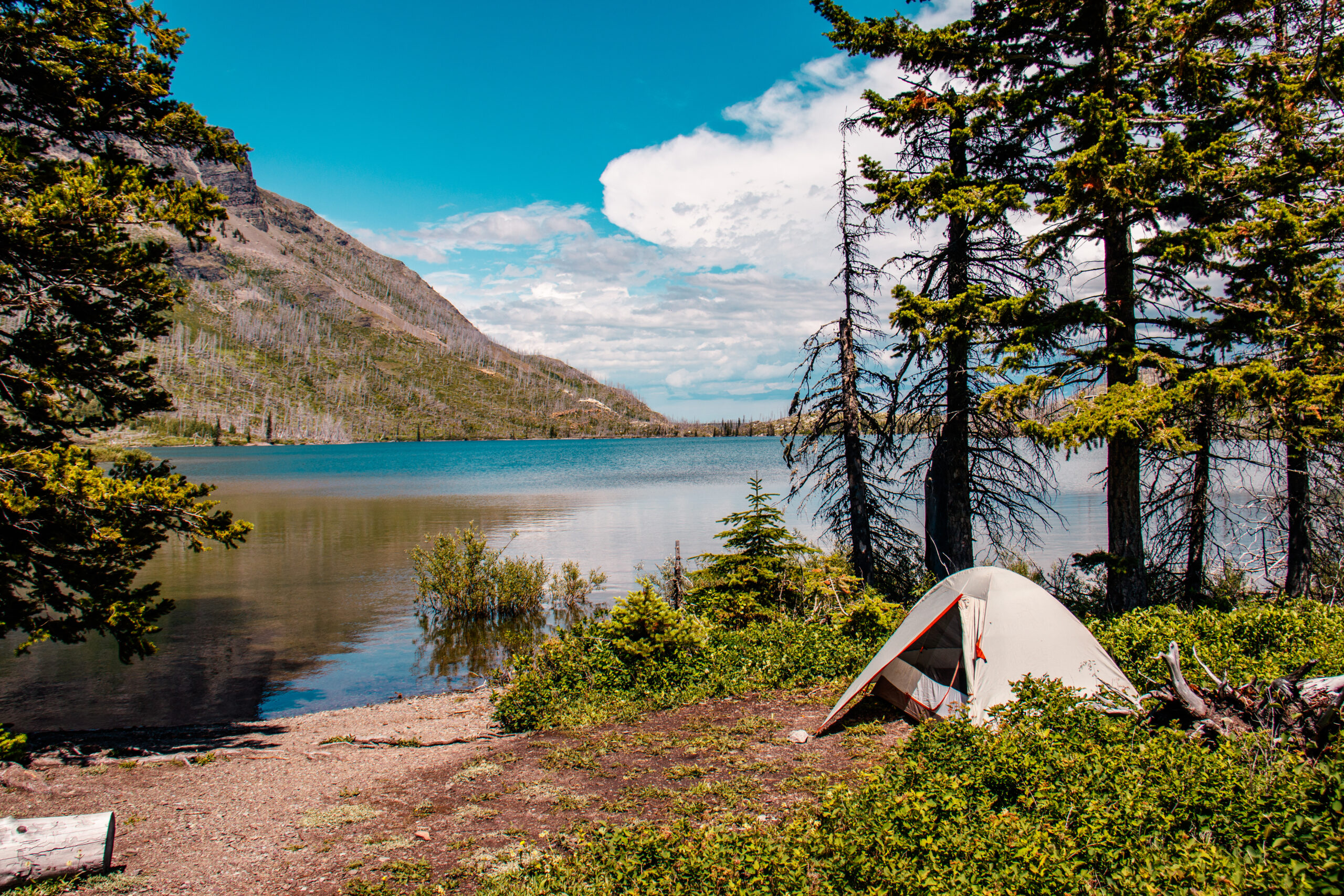 Photograph of a tent alongside a mountain lake on a mostly clear day in Glacier National Park. 