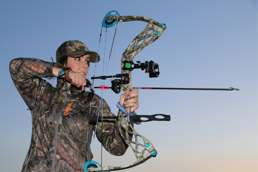 woman hunter poses for a photo while drawing back on a camo compound bow with teal highlights