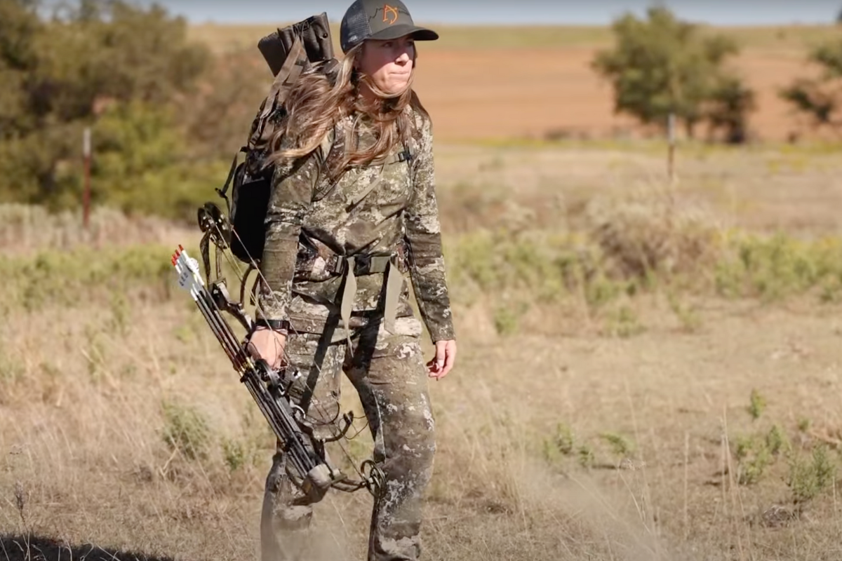 a camo-clad female bow hunter walks through tall grass