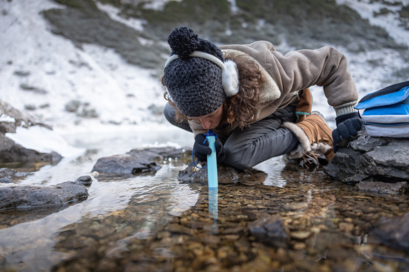 Adult Woman Using Filtrating Drinking Straw To Drink Water From a Lake in Winter.