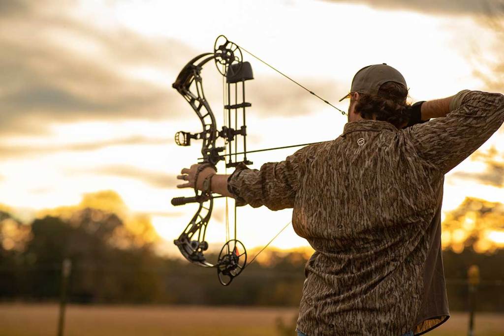 A hunter in camo aims a hunting bow with a sunset in the background