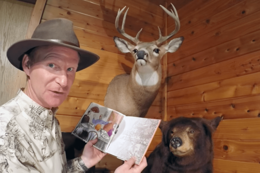 screenshot of video of author kevin lovegreen reading from children's book, wood paneled wall behind him with mounted heads of a buck and a bear
