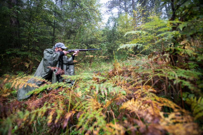 Portrait of hunter crouching in the forest and aiming with rifle.