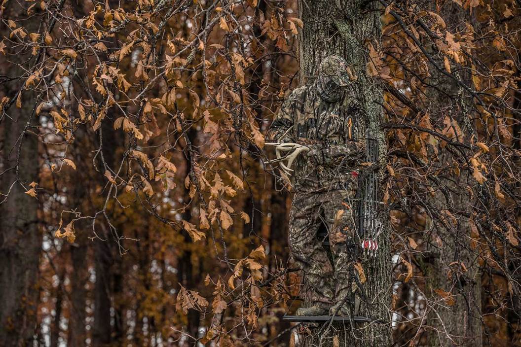 A hunter in full camo stands in a tree while holding deer antlers