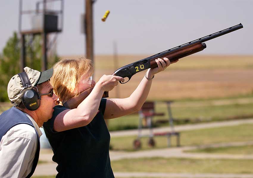 Woman aims a shotgun while an instructor looks on at a shooting range.