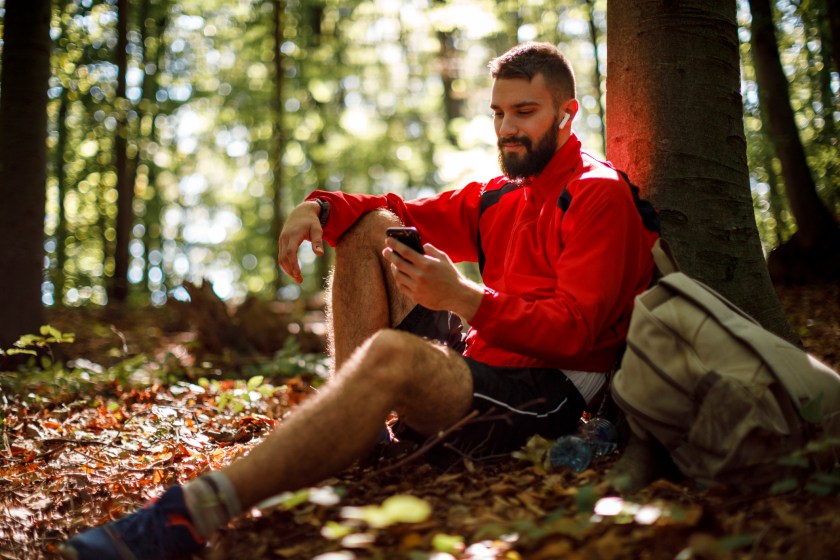 Portrait of young smiling man with bluetooth headphones using mobile phone in forest