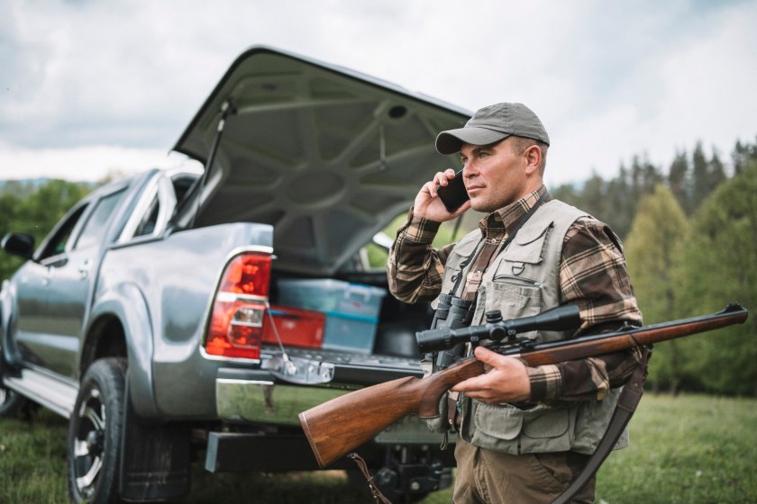 A hunter on a phone and holding his gun in front of his pickup truck.