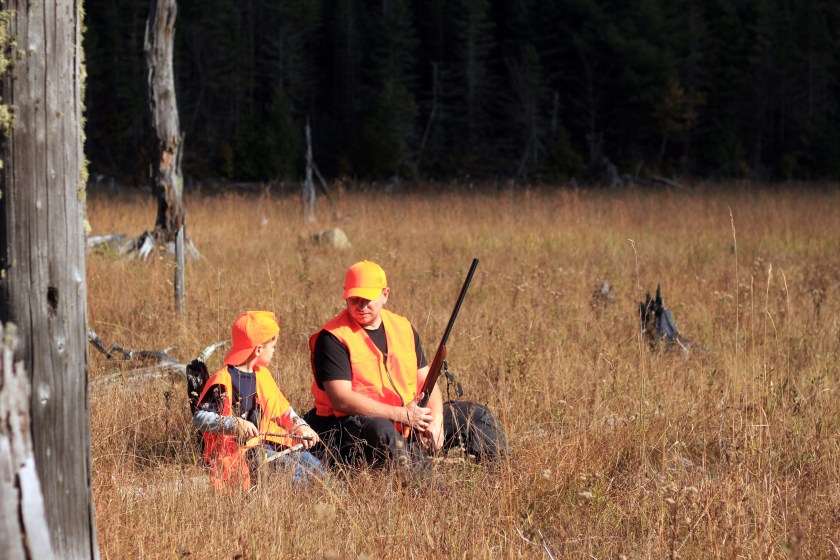A father and son on a hunt together, sitting in a field, wearing blaze orange vests.